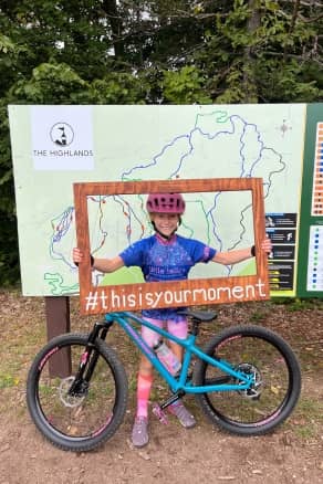 A child in bike gear holding a sign next to The Highlands Bike Park Trail Map at The Higlands