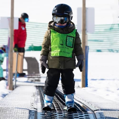 A small child in ski gear going up the magic carpet at The Highlands